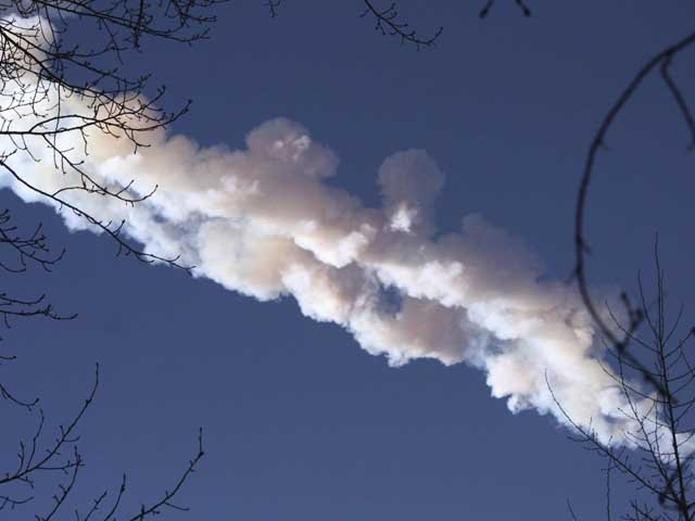 Smuga białego dymu po meteorze na błękitnym niebie, widziana zza gałęzi drzew